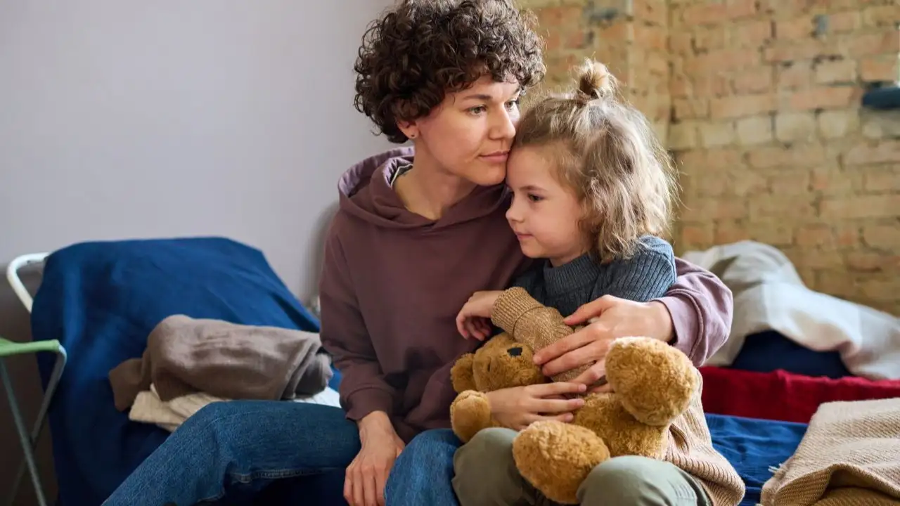 Young woman giving hug to her cute little son with brown soft teddybear while both sitting on sleeping place prepared for refugees