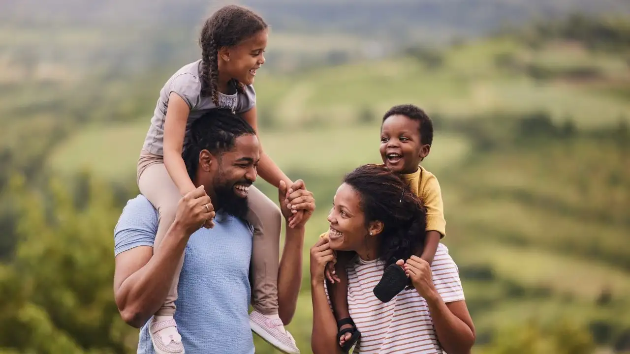 Happy parents carrying their small kids on shoulders in nature. -