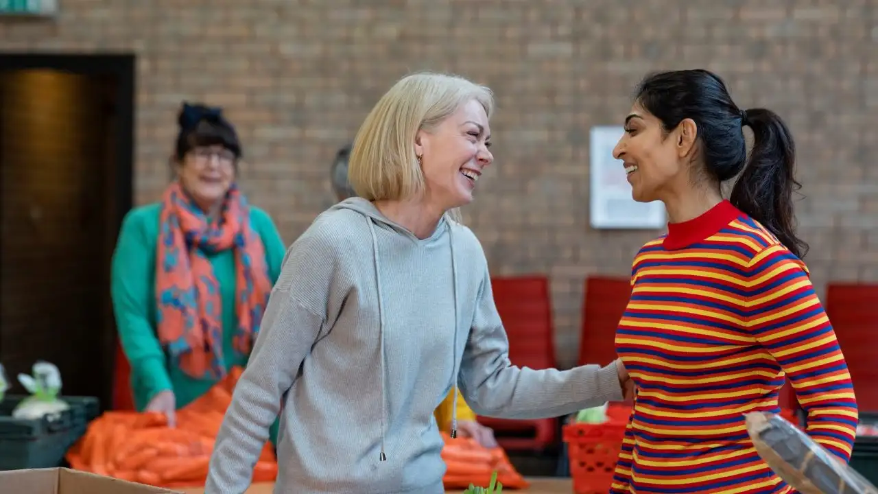 Group of women volunteers organizing food donations onto tables at a food bank.. They are looking at each other, laughing.